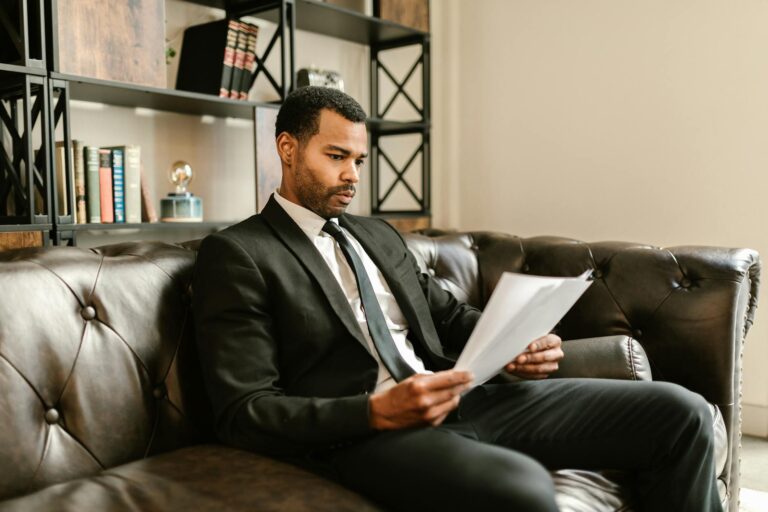 Businessman in suit reading papers on a leather sofa in a modern office setting.