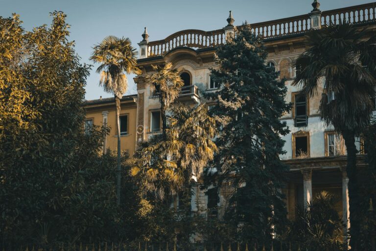 A mysterious abandoned villa surrounded by lush trees in a warm sunset light.