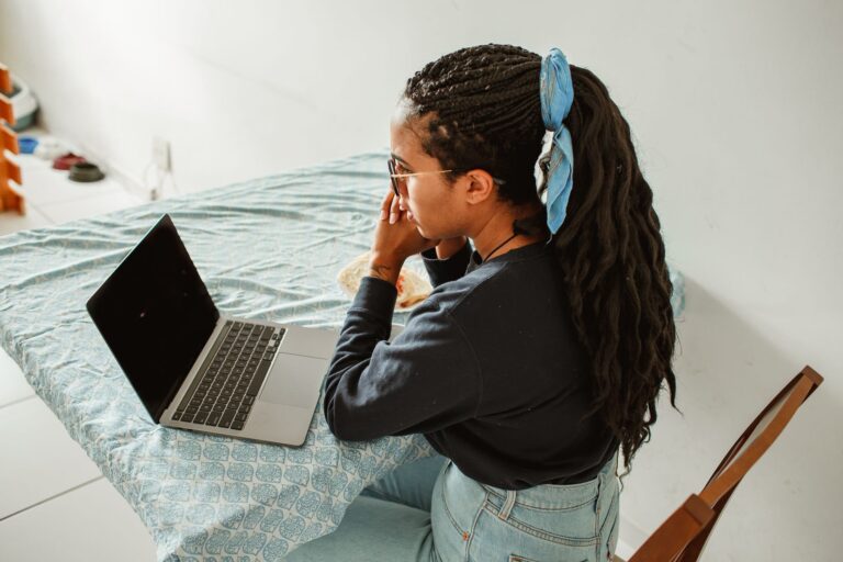 A young woman with braids and glasses studying on her laptop at a home table.