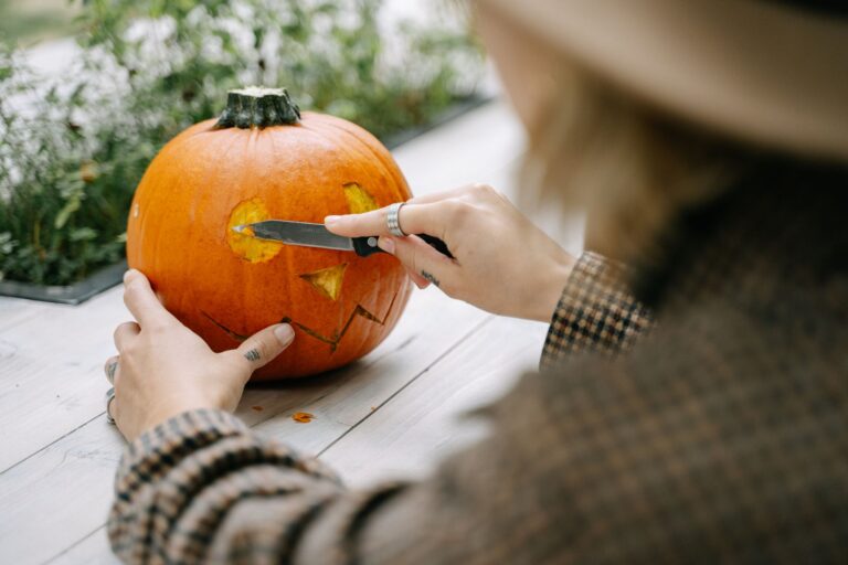 A woman carves a Halloween pumpkin with a knife on a wooden table outdoors.