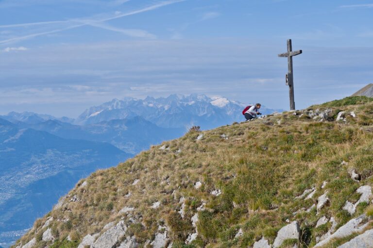 A hiker pauses at a mountain peak marked by a cross, overlooking stunning Alpine views.