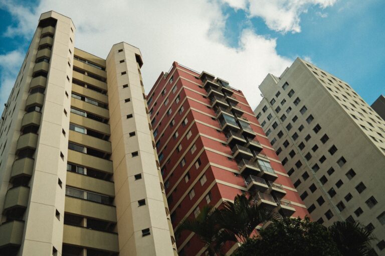 A low-angle view of three high-rise buildings with diverse architecture under a clear blue sky, highlighting urban life.